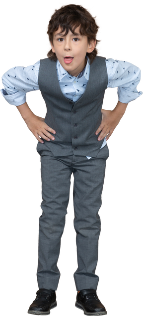 Вид спереди мальчика в сером костюме, позирующего с руками на бедрах и показывающим язык