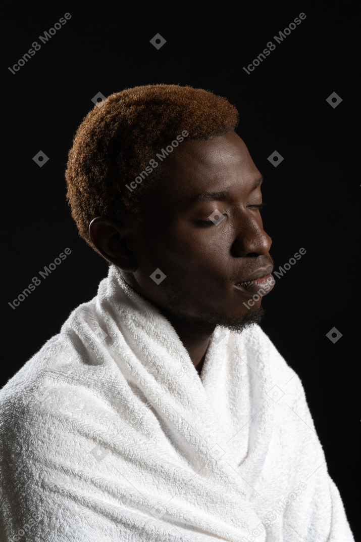 Plano lateral de un hombre sentado pacíficamente envuelto en una toalla con los ojos cerrados