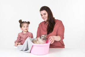 어머니와 그녀의 어린 딸, 빨간색과 분홍색 옷을 입고 가족 고양이와 함께 저녁 식사 테이블에 앉아