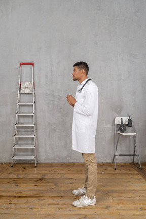 Vista lateral de un joven médico de pie en una habitación con escalera y silla explicando algo