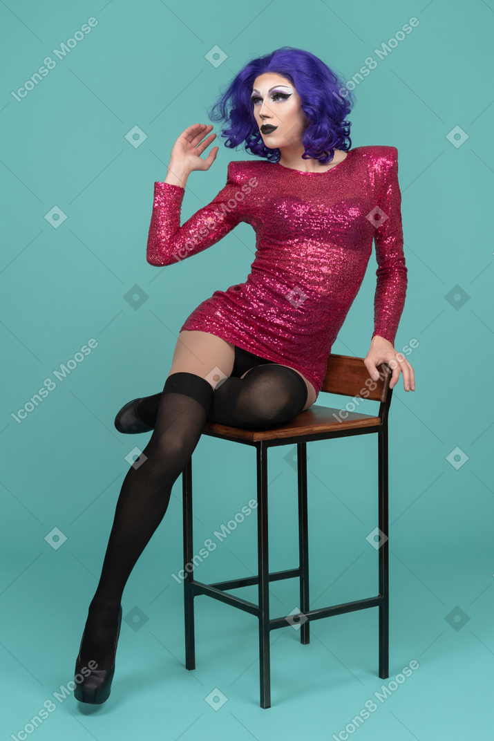 Drag queen flipping hair back & raising hand while sitting on a stool