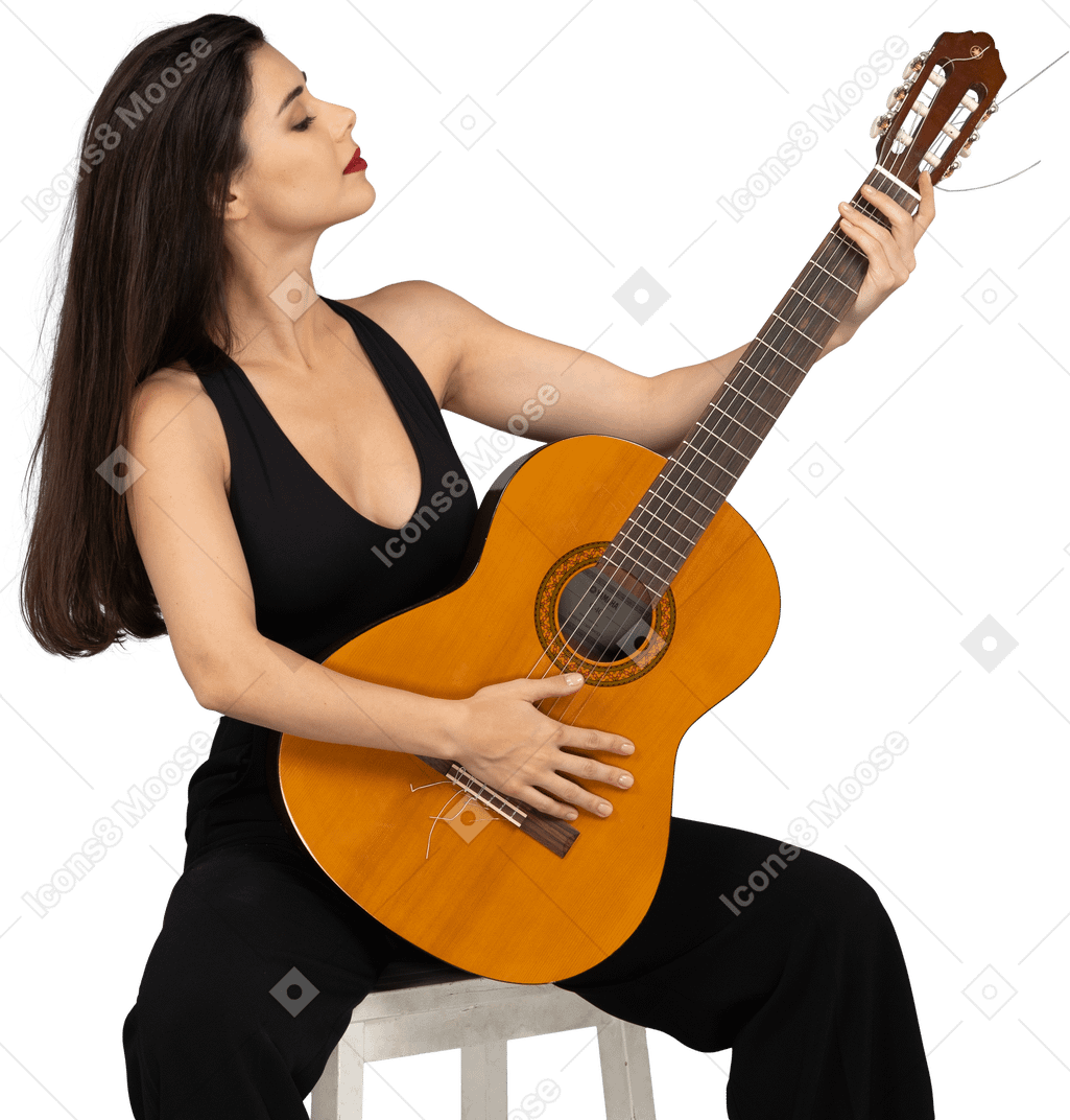 Vista frontal de uma jovem sentada de terno preto olhando orgulhosamente para seu violão