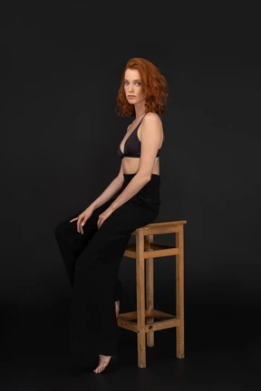 A side view of the sexy elegant woman, sitting on the wooden chair, holding her hands on knees and looking to the camera