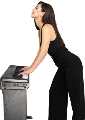 Vista lateral de uma jovem sorridente de terno preto tocando piano enquanto levanta a cabeça
