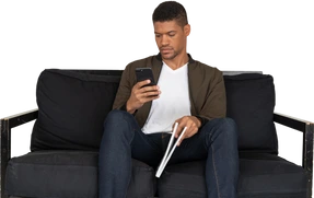 Front view of young man sitting on a sofa holding a magazine while using phone