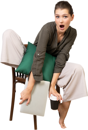 Vue de face d'une jeune femme choquée assise sur une chaise et tenant son ordinateur portable et touchant une tasse de café