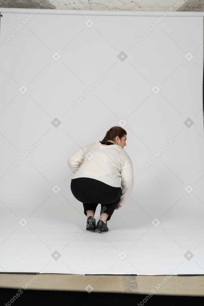 Вид сзади на сидящую на корточках женщину больших размеров в белом свитере