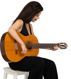 Vista de tres cuartos de una joven sentada en traje negro tocando la guitarra y mirando hacia abajo