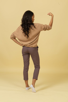 Back view of a strong dark-skinned young female raising hand while putting hand on hip