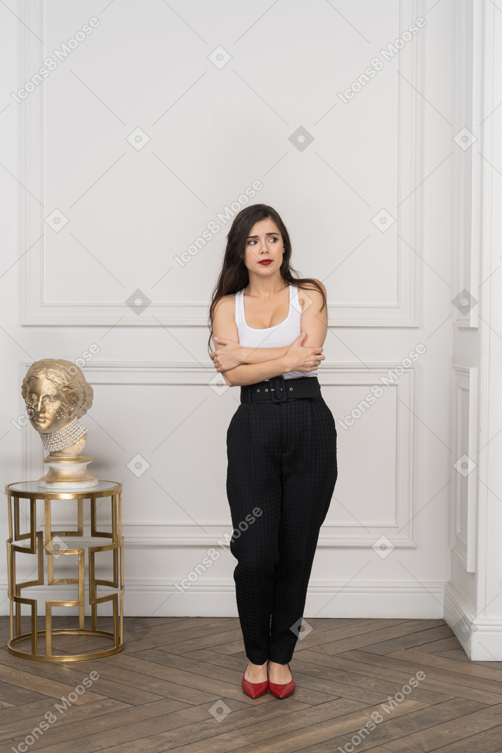 Corpo inteiro de uma jovem mulher cruzando as mãos em pé perto de uma escultura grega dourada
