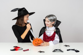 Boy in vampire costume and mom in witch costume carving a pumpkin