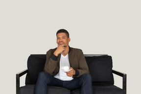 Front view of a young thoughtful man sitting on a sofa with a cup of coffee