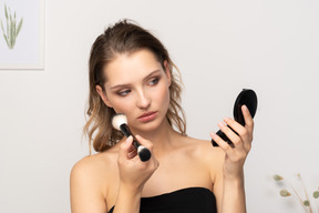 Front view of a young woman applying face powder while holding a mirror