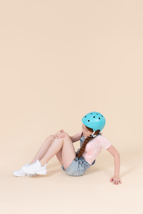 Teenager girl in blue helmet sitting on the floor and seems like got her knee hurt