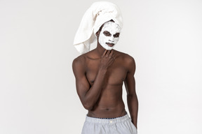 A young black man in grey trousers and a towel on his head going about his skin care routine