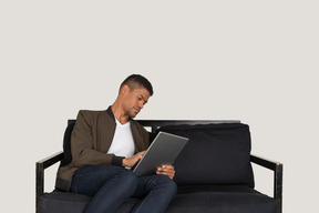 Vorderansicht eines jungen mannes, der auf einem sofa sitzt, während er ein tablet hält