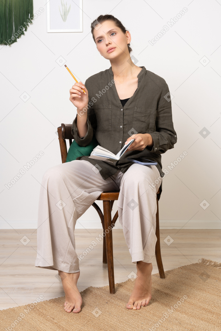 연필과 공책으로 의자에 앉아 집 옷을 입고 사려 깊은 젊은 여자의 전면보기
