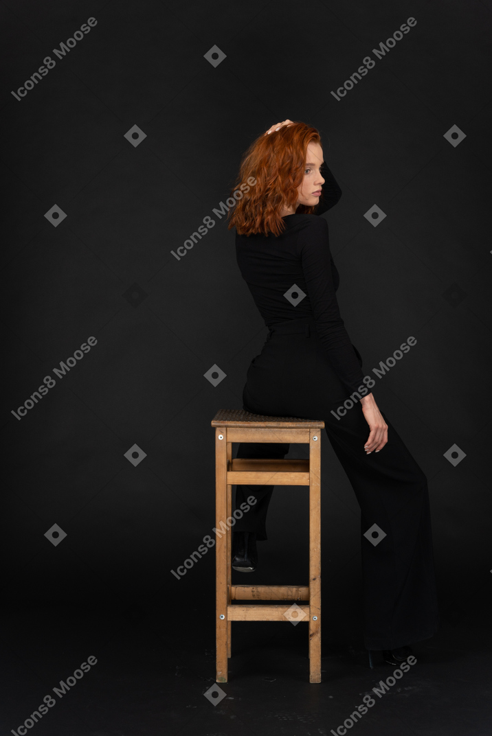 Una vista lateral trasera de la hermosa joven sentada en la silla alta de madera y ajustando su cabello
