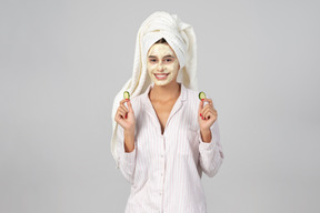 Ragazza con i capelli avvolti in un asciugamano e maschera sul viso in possesso di fette di cetriolo