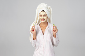 Girl with hair wrapped in towel and mask on her face holding cucumber slices