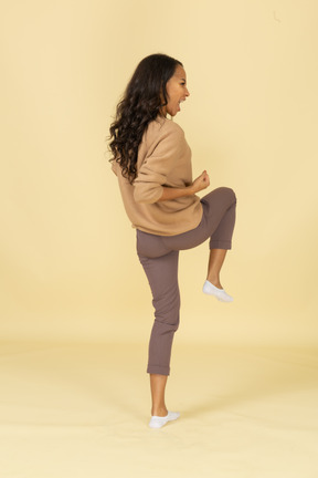 Vista posterior de tres cuartos de una joven mujer fresca de piel oscura levantando la pierna y apretando el puño