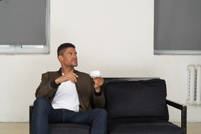 Vista de três quartos de um jovem sonhador sentado em um sofá com uma xícara de café