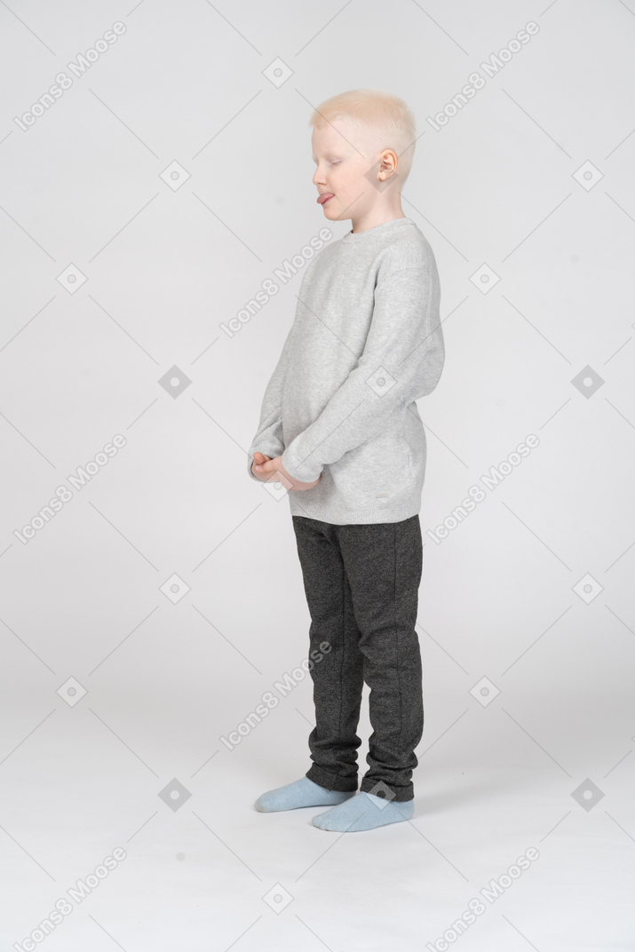 Three-quarter view of a kid boy in casual clothes showing tongue with his eyes closed putting hands together