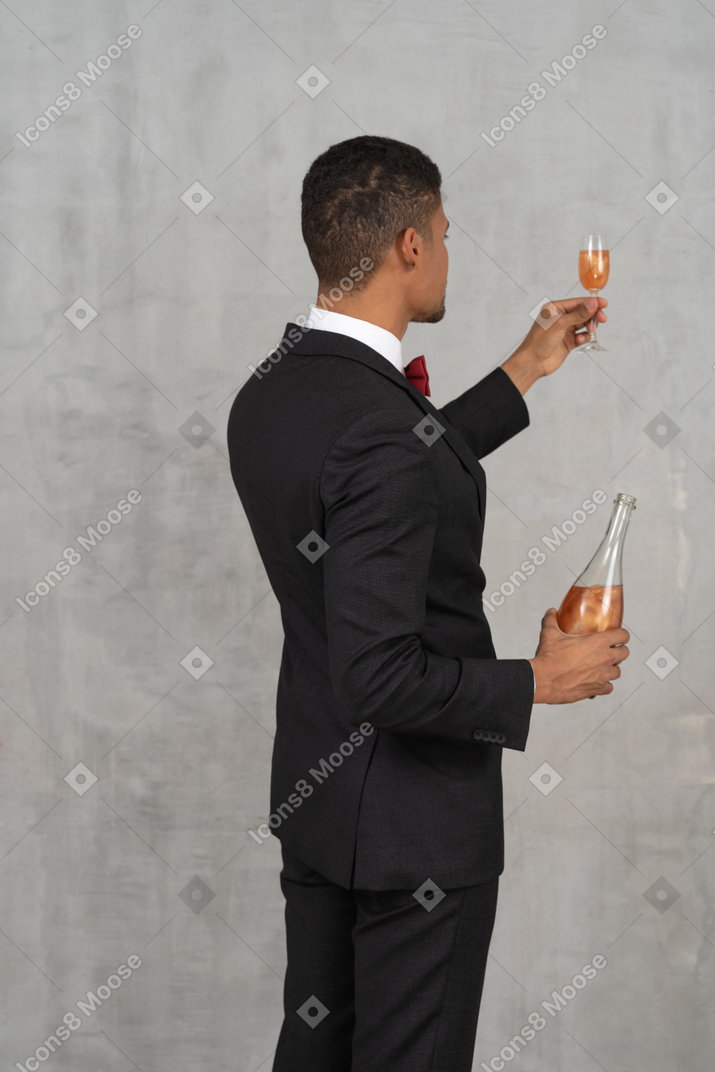 Punto di vista posteriore del giovane che tiene una bottiglia di liquore e un bicchiere di flauto
