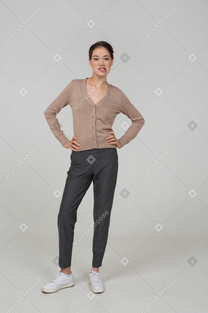 Front view of a young lady in pullover and pants putting hands on hips and clenching teeth