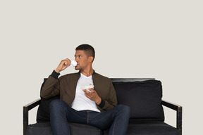 Front view of a young dreaming man sitting on a sofa while drinking coffee