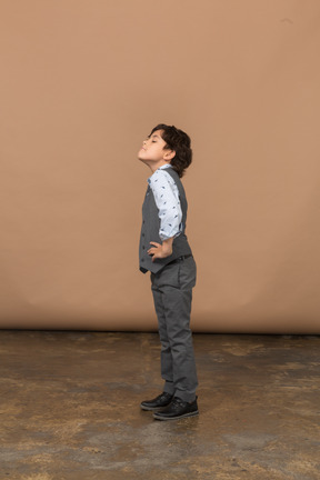 Seitenansicht eines süßen jungen im grauen anzug, der mit den händen auf den hüften steht und nach oben schaut