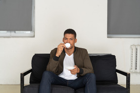 Front view of a young dreaming man sitting on a sofa while drinking coffee