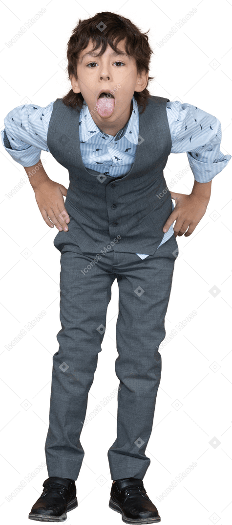 Vista frontal de um menino de terno cinza posando com as mãos nos quadris e mostrando a língua