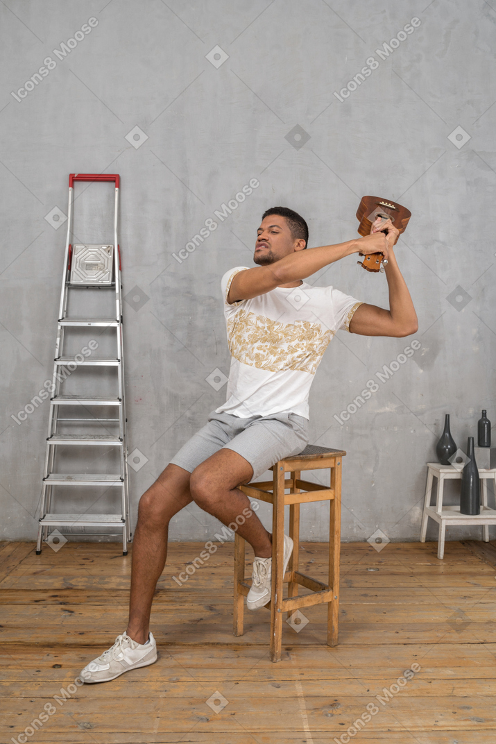 Three-quarter view of a man on a stool swinging an ukulele angrily