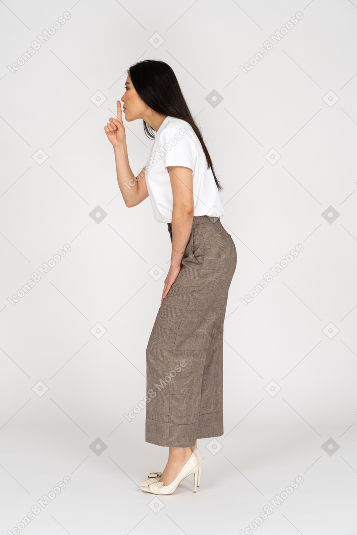 Vista lateral de uma jovem senhora de calça e camiseta mostrando um gesto de silêncio