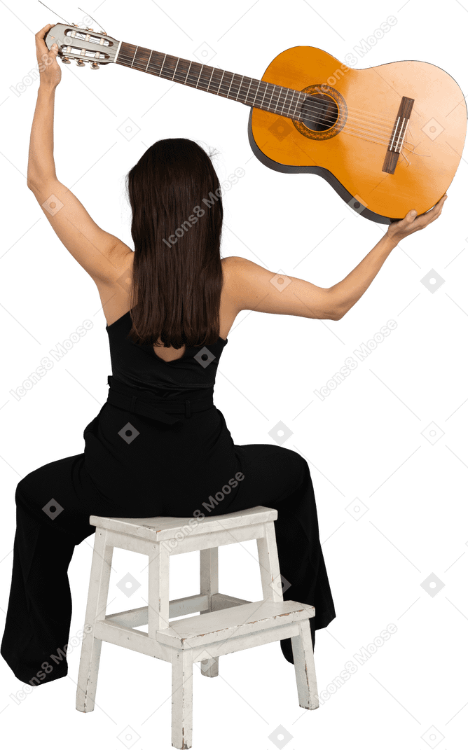 Вид сзади молодой женщины в черном костюме, держащей гитару над головой и сидящей на стуле