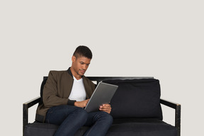 Front view of a young man sitting on a sofa while holding tablet