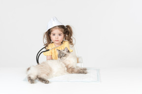 Little kid girl with stethoscope and wearing medical hat curing a cat