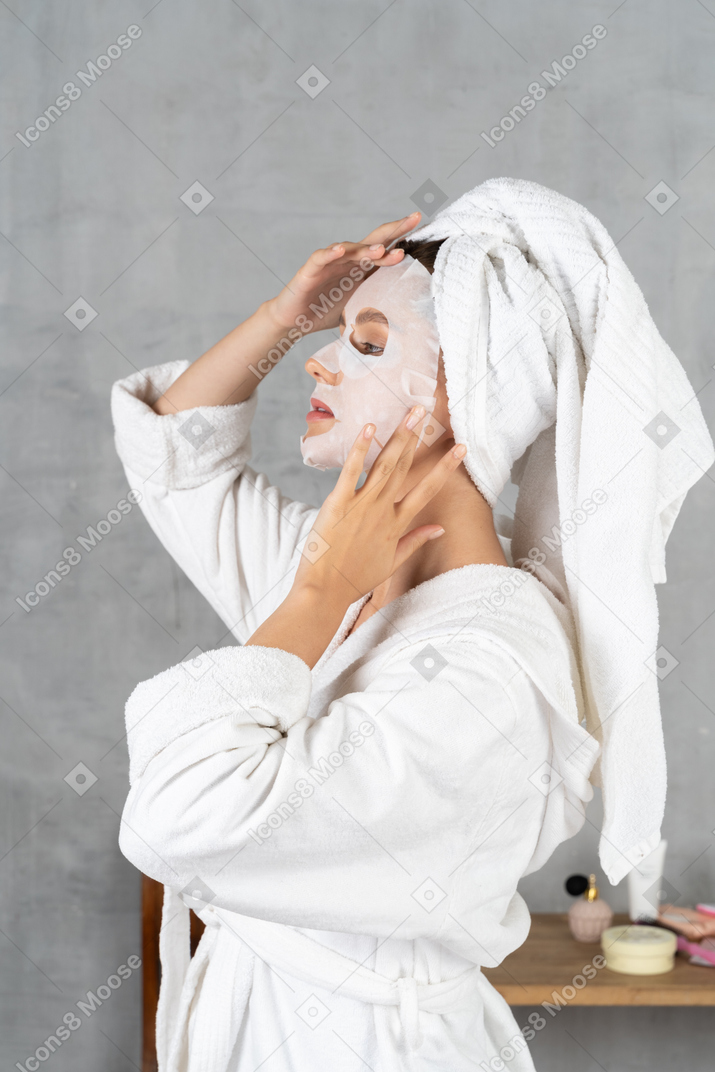 Side view of a woman in bathrobe applying a sheet mask