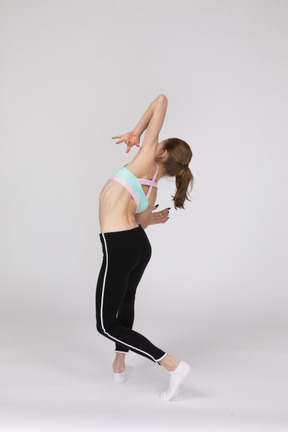 Three-quarter back view of a graceful teen girl in sportswear raising hands and tilting