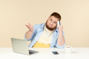 Dudoso joven con sobrepeso sentado en el escritorio de la oficina y haciendo compras en línea