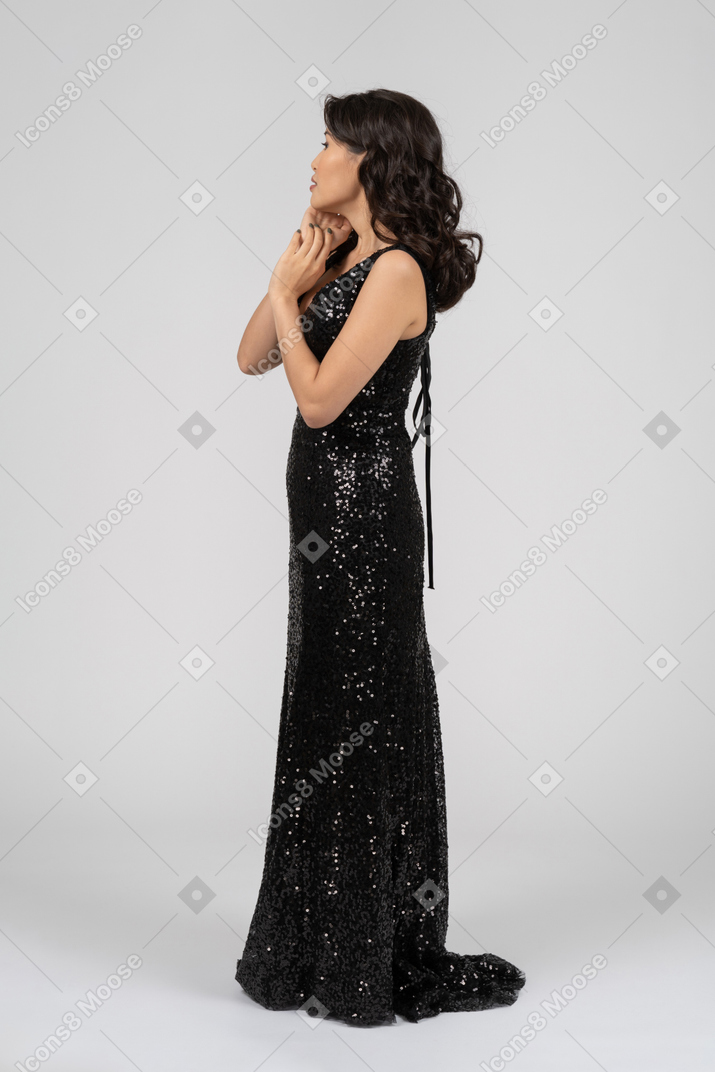 Женщина в черном вечернем платье стоит в профиль на камеру