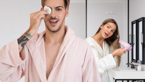 Young man holding a cotton pad standing next to a woman drying her hair in bathroom
