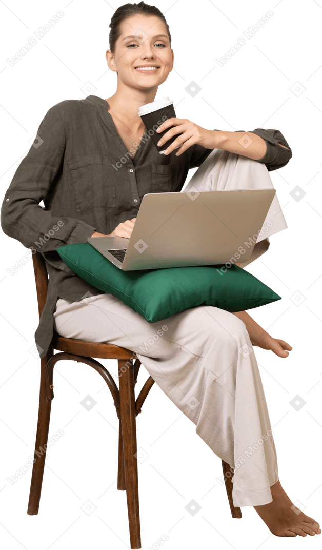 椅子に座って、彼女のラップトップとコーヒーカップを保持している笑顔の若い女性の正面図