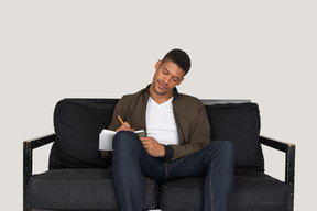 Vue de face du jeune homme assis sur un canapé et prenant des notes