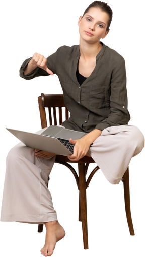 Vista frontal de una mujer joven disgustada sentada en una silla con una computadora portátil y mostrando el pulgar hacia abajo