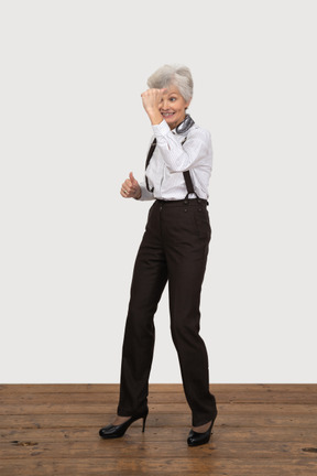 Трехчетвертный вид улыбающейся старушки в офисной одежде, поднимающей руку
