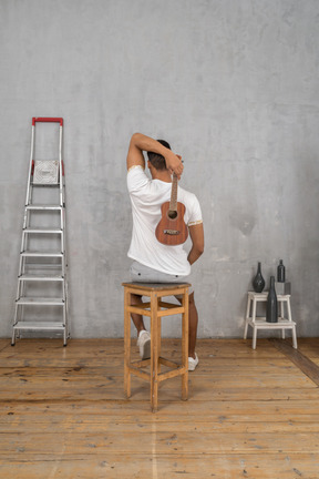 Rückansicht eines mannes auf einem hocker, der hinter seinem rücken eine ukulele hält