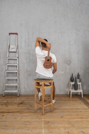 Vista trasera de un hombre en un taburete sosteniendo un ukelele detrás de su espalda