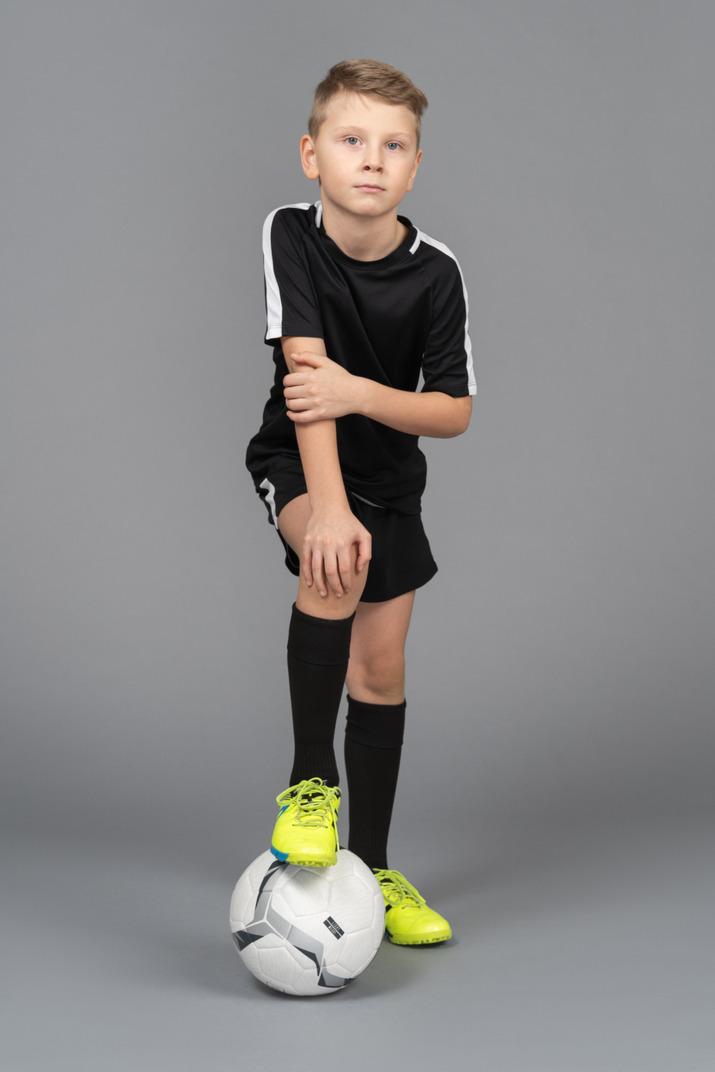 Front view of a child boy in football uniform putting his foot on ball